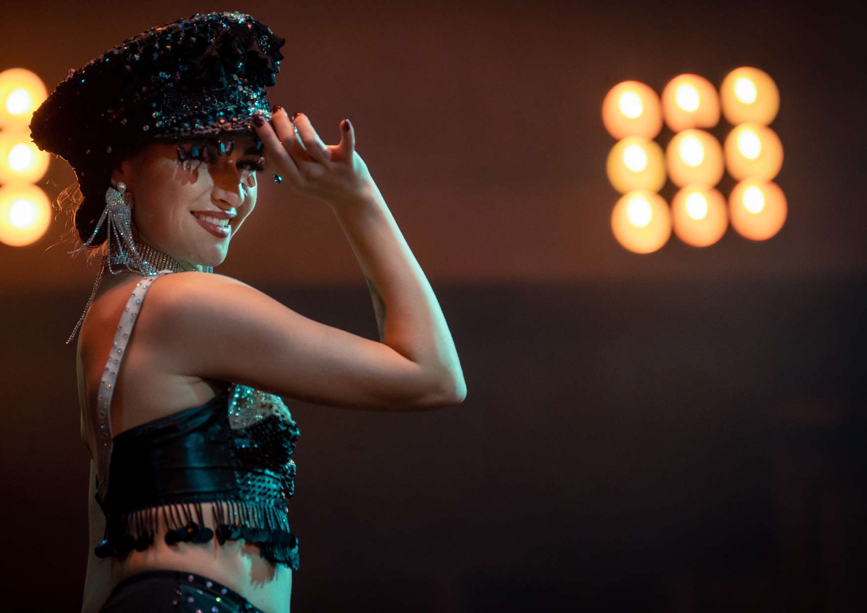 A cabaret performer in a black crystal costume and glittering cap, with warm golden stage lights